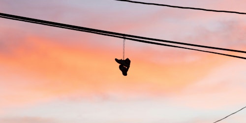 A pair of trainers or sneakers hangs over a telephone wire by the laces against a dawn or dusk sky A pair of trainers or sneakers hangs over a telephone wire by the laces against a dawn or dusk sky