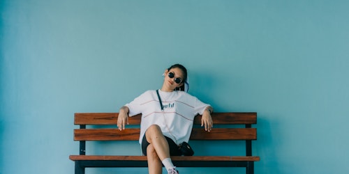 Young adult woman sat on bench in baggy t-shirt & sunglasses against blue background Young adult woman sat on bench in baggy t-shirt & sunglasses against blue background