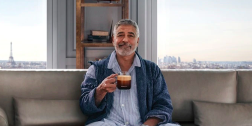 George Clooney sits on a sofa holding a cup of black coffee George Clooney sits on a sofa holding a cup of black coffee